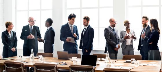 Gruppo di uomi e donne in abiti formali che discute in piedi in una sala riunioni luminosa.